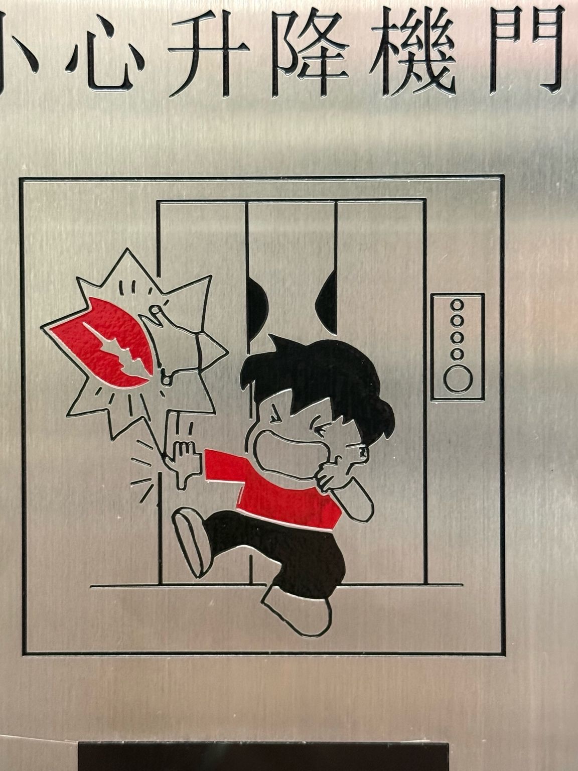 Photo of a Hong Kong lift warning plaque reading Mind the lift door in Chinese, with a cartoon boy crying as his hand gets caught in the closing door.